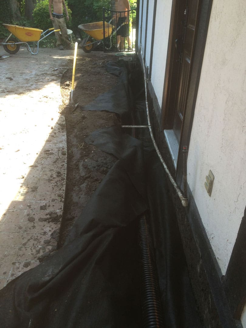 A trench dug alongside a house with black plastic sheeting laid inside.